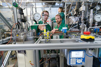 Foto: Drei Personen in grünen Kitteln arbeiten in einem Labor an einer komplexen technischen Anlage mit Druckanzeigen und Schläuchen. Ein Display zeigt "7.02" und "38.2°C". Die Umgebung ist industriell mit zahlreichen Messgeräten und Kabeln.