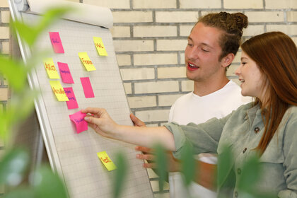 Foto: Zwei Personen stehen vor einem Flipchart mit bunten Haftnotizen. Eine Person zeigt auf eine der Notizen. Der Hintergrund besteht aus einer Ziegelwand.