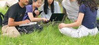 Studierende sitzen unter einem Baum im Gras und schauen auf einen Laptop.