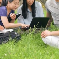 Studierende sitzen unter einem Baum im Gras und schauen auf einen Laptop.