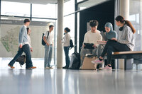 Foto: Mehrere Personen sitzen und stehen in einem modernen Universitätsgebäude. Einige sitzen auf einer Bank mit Laptops und Tablets, während andere im Hintergrund gehen. Eine Karte der Universität Hohenheim ist an der Wand sichtbar.