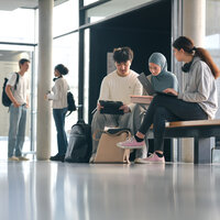 Foto: Mehrere Personen sitzen und stehen in einem modernen Universitätsgebäude. Einige sitzen auf einer Bank mit Laptops und Tablets, während andere im Hintergrund gehen. Eine Karte der Universität Hohenheim ist an der Wand sichtbar.