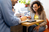 Foto: Zwei Personen sitzen an einem Tisch. Eine Person schreibt in ein Notizbuch, während die andere ein Tablet benutzt. Auf dem Tisch liegen Bücher, ein Getränk und ein Taschenrechner.
