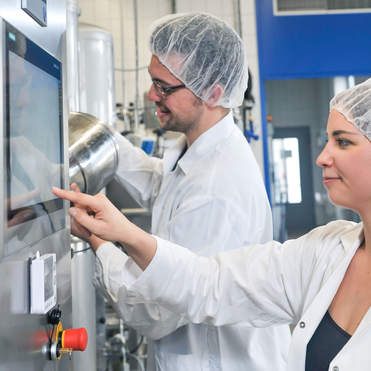 Foto: Zwei Personen in weißen Laborkitteln und Haarnetzen bedienen eine Maschine mit einem Touchscreen in einem Labor. Eine Person zeigt auf den Bildschirm, während die andere an einem Behälter arbeitet. Im Hintergrund sind industrielle Geräte sichtbar.
