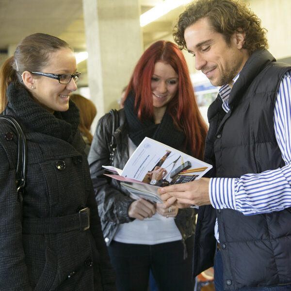 Foto: Drei Personen stehen in einem Innenraum und betrachten gemeinsam Broschüren. Eine Person hält eine Broschüre mit Bildern und Text, während die anderen aufmerksam zuhören. Die Umgebung wirkt wie ein öffentlicher oder akademischer Raum.