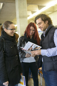 Foto: Drei Personen stehen in einem Innenraum und betrachten gemeinsam Broschüren. Eine Person hält eine Broschüre mit Bildern und Text, während die anderen aufmerksam zuhören. Die Umgebung wirkt wie ein öffentlicher oder akademischer Raum.