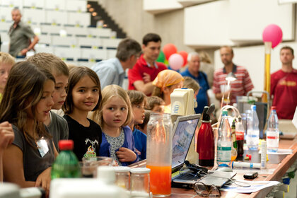 Foto: Eine Gruppe von Personen, darunter Kinder, steht an einem Tisch mit wissenschaftlichen Geräten und einem Laptop. Im Hintergrund sind weitere Personen und bunte Luftballons zu sehen.