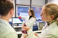 Foto: Drei Personen in Laborkitteln arbeiten in einem Labor. Eine Person hält einen Kolben mit roter Flüssigkeit, während eine andere Person eine Schale mit roten Zwiebeln hält. Im Hintergrund ist ein Computerbildschirm sichtbar.