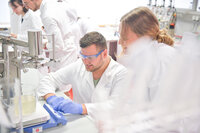 Foto: Mehrere Personen in weißen Laborkitteln arbeiten in einem Labor. Eine Person trägt blaue Handschuhe und bedient ein Gerät. Im Hintergrund sind weitere Laborgeräte sichtbar.