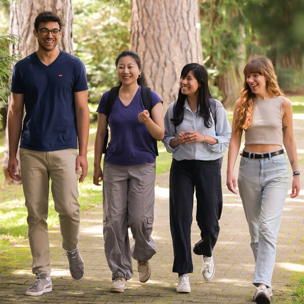 Foto: Vier Personen gehen auf einem gepflasterten Weg in einem Park, umgeben von hohen Bäumen und grüner Vegetation.