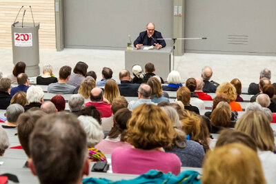 Foto: Ein Hörsaal mit vielen Personen, die einem Vortragenden zuhören. Der Redner sitzt an einem Tisch vor einer grauen Wand. Links steht ein Rednerpult mit der Aufschrift "200". Die Sitzreihen sind mit Menschen gefüllt, die aufmerksam nach vorne schauen.