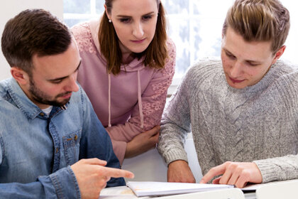 Foto: Drei Personen sitzen an einem Tisch und arbeiten gemeinsam an einem Dokument. Eine Person zeigt auf das Dokument, während die anderen beiden aufmerksam schauen. Die Umgebung wirkt wie ein heller Raum mit weißen Wänden.