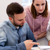Foto: Drei Personen sitzen an einem Tisch und arbeiten gemeinsam an einem Dokument. Eine Person zeigt auf das Dokument, während die anderen beiden aufmerksam schauen. Die Umgebung wirkt wie ein heller Raum mit weißen Wänden.