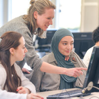 Lehrende zeigt drei Studierenden etwas am Bildschirm in einem Computerraum.