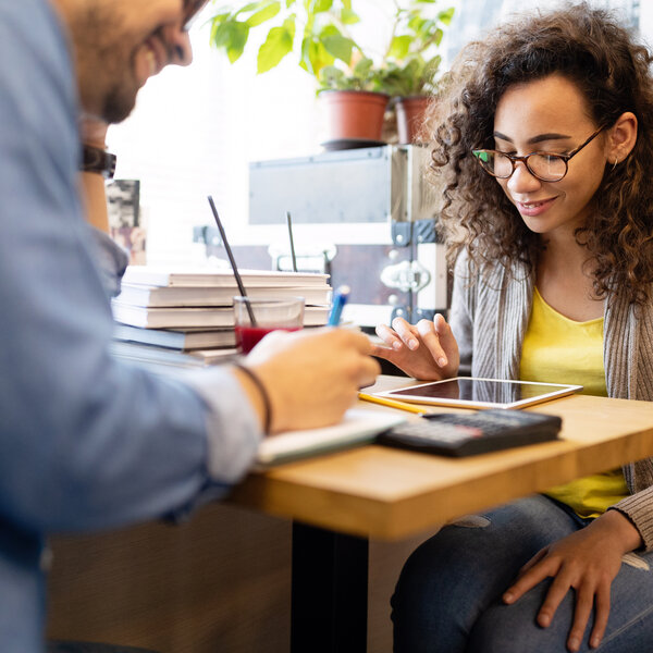 Foto: Zwei Personen sitzen an einem Tisch. Eine Person schreibt in ein Notizbuch, während die andere ein Tablet benutzt. Auf dem Tisch liegen Bücher, ein Getränk und ein Taschenrechner.