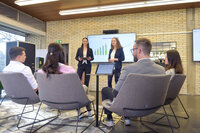 Foto: Zwei Personen in Anzügen präsentieren vor einem Publikum. Auf einem Bildschirm hinter ihnen ist eine Grafik mit dem Titel 'BREAK-EVEN-ANALYSE' zu sehen. Mehrere Personen sitzen im Vordergrund und hören zu.