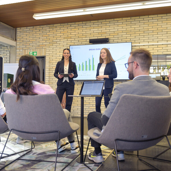 Foto: Zwei Personen in Anzügen präsentieren vor einem Publikum. Auf einem Bildschirm hinter ihnen ist eine Grafik mit dem Titel 'BREAK-EVEN-ANALYSE' zu sehen. Mehrere Personen sitzen im Vordergrund und hören zu.
