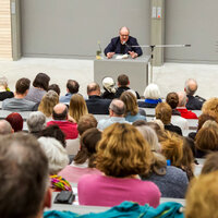 Foto: Ein Hörsaal mit vielen Personen, die einem Vortragenden zuhören. Der Redner sitzt an einem Tisch vor einer grauen Wand. Links steht ein Rednerpult mit der Aufschrift "200". Die Sitzreihen sind mit Menschen gefüllt, die aufmerksam nach vorne schauen.