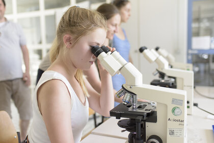 Foto: Mehrere Personen in einem Labor nutzen Mikroskope. Ein Mikroskop ist mit 'Axiostar plus' beschriftet. Die Umgebung wirkt akademisch.
