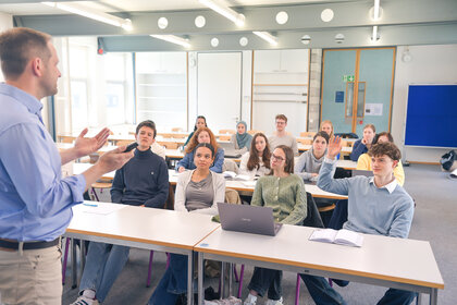 Foto: Eine Person steht vor einem Klassenzimmer und spricht zu mehreren Personen, die an Tischen sitzen. Einige Personen haben Laptops und Notizbücher vor sich. Eine Person hebt die Hand.