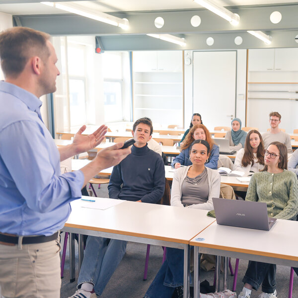 Foto: Eine Person steht vor einem Klassenzimmer und spricht zu mehreren Personen, die an Tischen sitzen. Einige Personen haben Laptops und Notizbücher vor sich. Eine Person hebt die Hand.