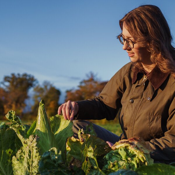 Foto: Eine Person in einem braunen Mantel sitzt auf einem Feld und untersucht große grüne Blätter. Im Hintergrund sind Bäume und ein klarer Himmel sichtbar.