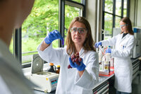 Studentin im Laborkittel hält Trauben und ein Reagenzglas, zweite Person arbeitet im Hintergrund.