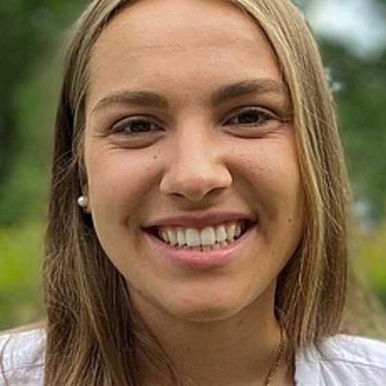 Foto: Eine Person mit langen Haaren steht im Freien vor einer grünen Hecke. Der Hintergrund zeigt Bäume und einen bewölkten Himmel. Die Person trägt ein weißes Oberteil.
