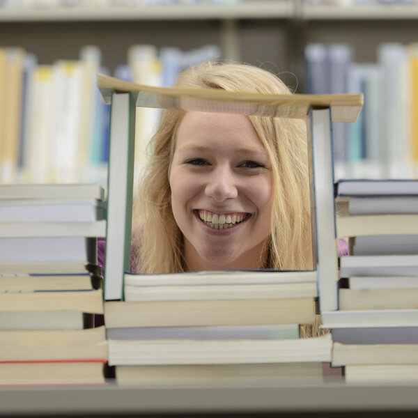 Foto: Eine Person sitzt hinter einem großen Stapel Bücher in einer Bibliothek. Im Hintergrund sind Bücherregale mit verschiedenen Büchern zu sehen.