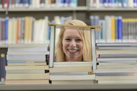 Foto: Eine Person sitzt hinter einem großen Stapel Bücher in einer Bibliothek. Im Hintergrund sind Bücherregale mit verschiedenen Büchern zu sehen.