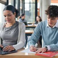 Foto: Zwei Personen sitzen an einem Tisch in einer Bibliothek. Eine Person arbeitet an einem Laptop, während die andere Notizen auf Papier macht. Im Hintergrund sind weitere Personen an Tischen sichtbar.