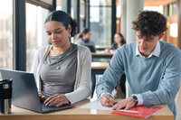 Foto: Zwei Personen sitzen an einem Tisch in einer Bibliothek. Eine Person arbeitet an einem Laptop, während die andere Notizen auf Papier macht. Im Hintergrund sind weitere Personen an Tischen sichtbar.