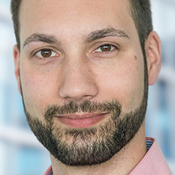 Foto: Eine Person in einem rosa Hemd steht vor einem modernen Gebäude mit Glasfassade. Der Hintergrund zeigt unscharfe Fensterstrukturen.