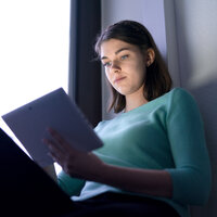 Foto: Eine Person sitzt in einem Raum und hält ein Tablet. Sie trägt einen türkisfarbenen Pullover. Das Licht im Raum ist gedämpft, und die Person scheint auf das Tablet zu schauen. Der Hintergrund zeigt eine Wand und ein Fenster.