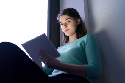 Foto: Eine Person sitzt in einem Raum und hält ein Tablet. Sie trägt einen türkisfarbenen Pullover. Das Licht im Raum ist gedämpft, und die Person scheint auf das Tablet zu schauen. Der Hintergrund zeigt eine Wand und ein Fenster.