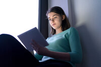 Foto: Eine Person sitzt in einem Raum und hält ein Tablet. Sie trägt einen türkisfarbenen Pullover. Das Licht im Raum ist gedämpft, und die Person scheint auf das Tablet zu schauen. Der Hintergrund zeigt eine Wand und ein Fenster.