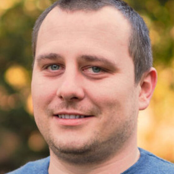 Foto: Eine Person steht im Freien vor einem unscharfen, herbstlichen Hintergrund. Die Person trägt ein blaues Oberteil. Die Umgebung zeigt warme, goldene Farbtöne, die auf herbstliches Laub hindeuten.