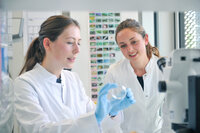 Zwei Studentinnen in weißen Laborkitteln betrachten gemeinsam eine Petrischale in einem Labor. 