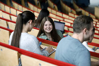 Foto: Mehrere Personen sitzen in einem Hörsaal mit Holzbänken und roten Geländern. Eine Person schreibt auf ein Tablet, während andere Personen Notizen machen. Ein Laptop ist im Hintergrund sichtbar.