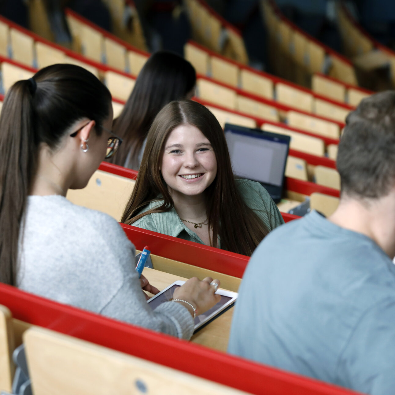 Foto: Mehrere Personen sitzen in einem Hörsaal mit Holzbänken und roten Geländern. Eine Person schreibt auf ein Tablet, während andere Personen Notizen machen. Ein Laptop ist im Hintergrund sichtbar.