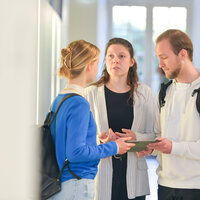Foto: Drei Personen stehen in einem hellen Raum und unterhalten sich. Eine Person trägt einen blauen Pullover und einen Rucksack, eine andere einen gestreiften Blazer, und die dritte eine weiße Kapuzenjacke mit einem Rucksack.