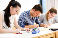 Foto: Drei Personen sitzen an einem Tisch in einem Klassenzimmer und schreiben auf Papier. Auf dem Tisch liegen ein Federmäppchen und ein Stift. Die Umgebung wirkt hell und modern.