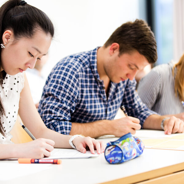 Foto: Drei Personen sitzen an einem Tisch in einem Klassenzimmer und schreiben auf Papier. Auf dem Tisch liegen ein Federmäppchen und ein Stift. Die Umgebung wirkt hell und modern.