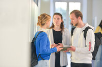 Foto: Drei Personen stehen in einem hellen Raum und unterhalten sich. Eine Person trägt einen blauen Pullover und einen Rucksack, eine andere einen gestreiften Blazer, und die dritte eine weiße Kapuzenjacke mit einem Rucksack.