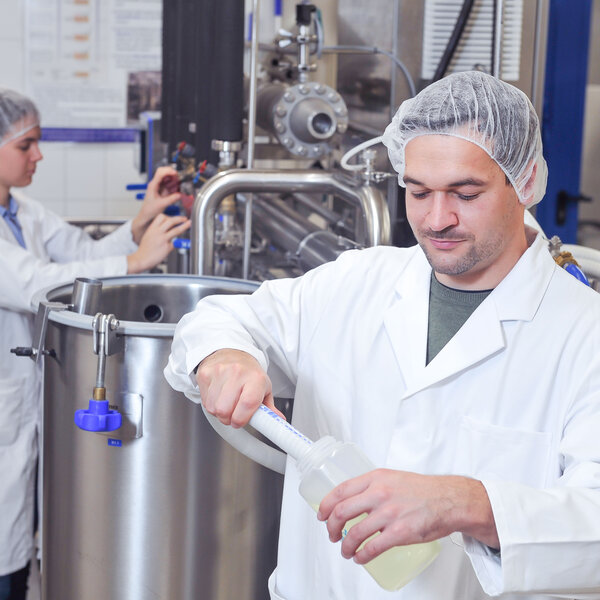 Foto: Zwei Personen in weißen Laborkitteln und Haarnetzen arbeiten in einem Labor. Eine Person hält eine Flasche und ein Werkzeug, während die andere an einem großen Edelstahlbehälter arbeitet. Die Umgebung ist industriell mit verschiedenen Maschinen und Rohren.