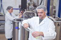 Foto: Zwei Personen in weißen Laborkitteln und Haarnetzen arbeiten in einem Labor. Eine Person hält eine Flasche und ein Werkzeug, während die andere an einem großen Edelstahlbehälter arbeitet. Die Umgebung ist industriell mit verschiedenen Maschinen und Rohren.