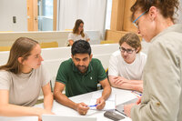 Foto: Mehrere Personen sitzen an einem Tisch in einem Klassenzimmer und arbeiten zusammen. Eine Person steht und scheint Anweisungen zu geben. Auf dem Tisch liegen Notizbücher, Stifte und ein Laptop. Im Hintergrund sind weitere Sitzreihen zu sehen.