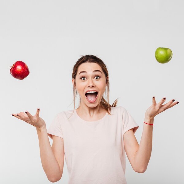 Foto: Eine Person in einem hellen T-Shirt jongliert mit einem roten und einem grünen Apfel vor einem neutralen Hintergrund. Die Arme sind seitlich ausgestreckt, während die Äpfel in der Luft schweben.