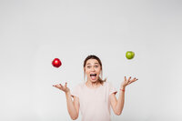 Foto: Eine Person in einem hellen T-Shirt jongliert mit einem roten und einem grünen Apfel vor einem neutralen Hintergrund. Die Arme sind seitlich ausgestreckt, während die Äpfel in der Luft schweben.