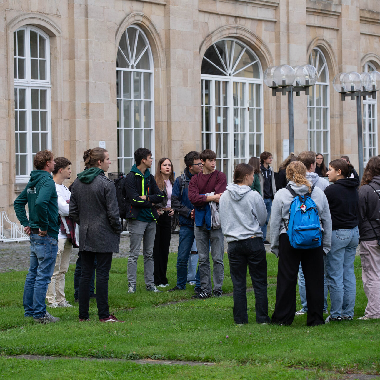 Foto: Eine Gruppe von Personen steht auf einem Rasen vor dem Schloss Hohenheim. Das Gebäude im Hintergrund hat große Fenster und klassische Architekturmerkmale. Die Personen tragen verschiedene Freizeitkleidung und scheinen in ein Gespräch vertieft zu sein.